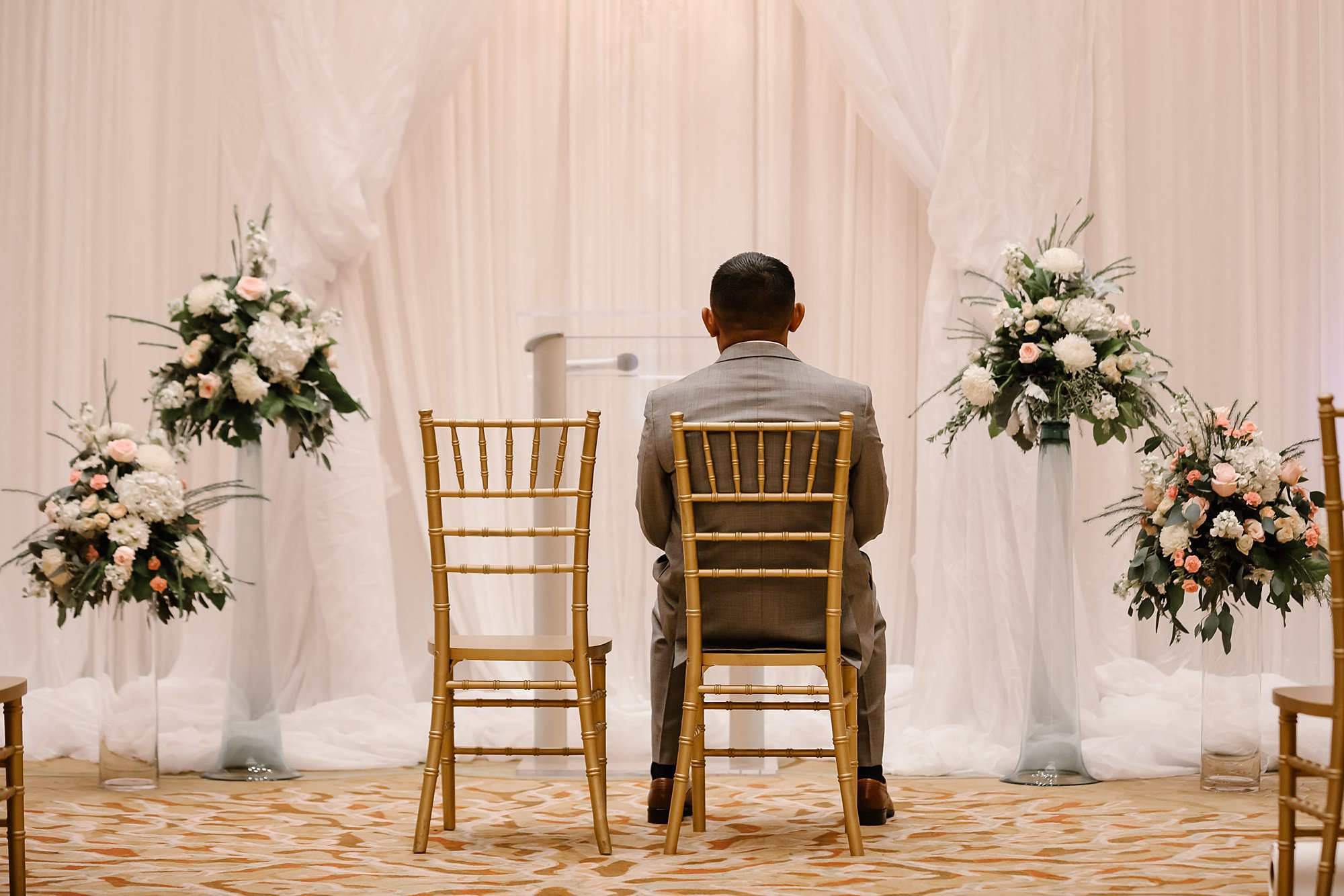 Groom sitting in a gold bamboo chair in front of the alter waiting for his bride to walk in