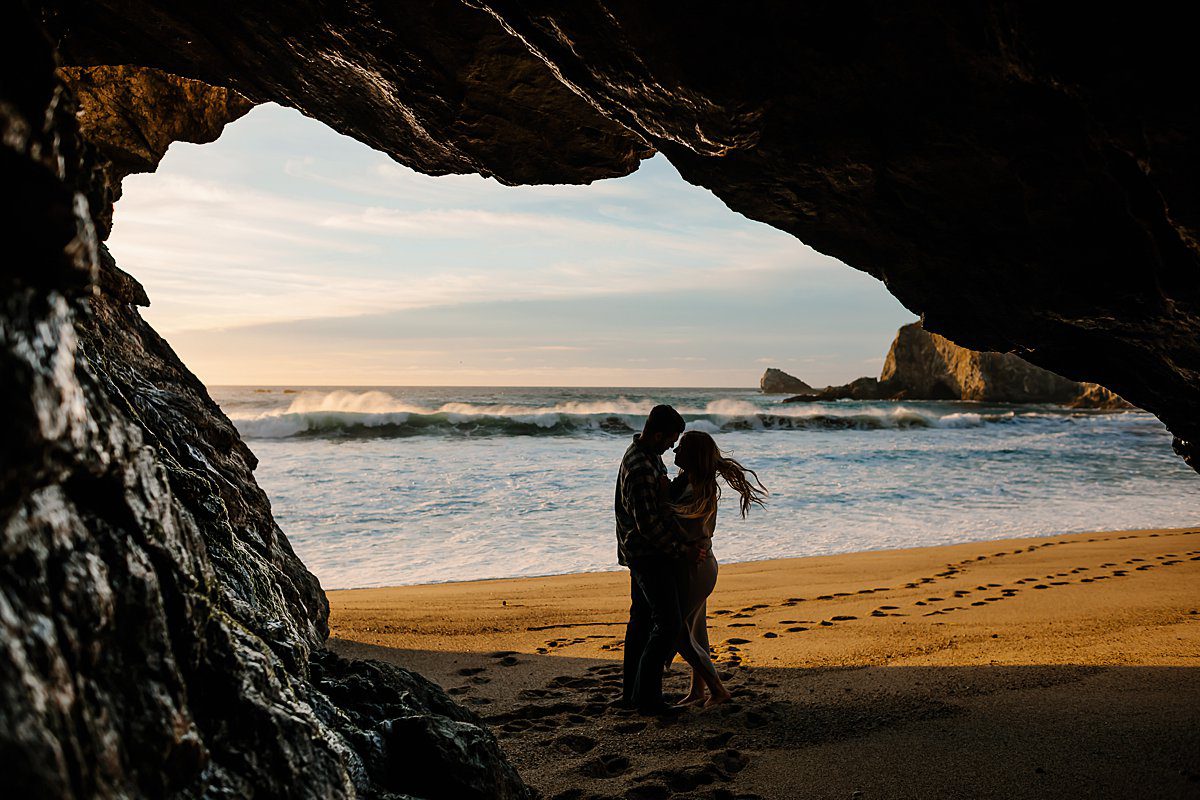 View from a cave, of a couple silhouetted in the sunset embracing on the beach in Big Sur under the Bixby Bridge.