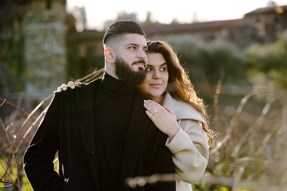 Close up of woman embracing her fiance from behind while standing in the winter vineyard in front of Castello di Amorosa