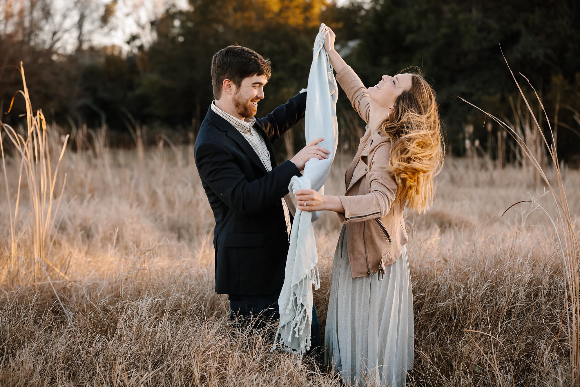 Megan and Turner laughing trying to unfurl a light blue blanket while in a field of tall dry grass