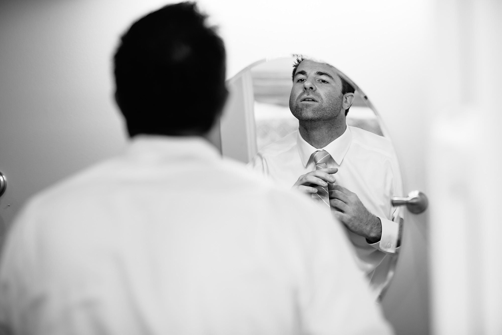 Black and white of groom adjusting tie in round bathroom mirror