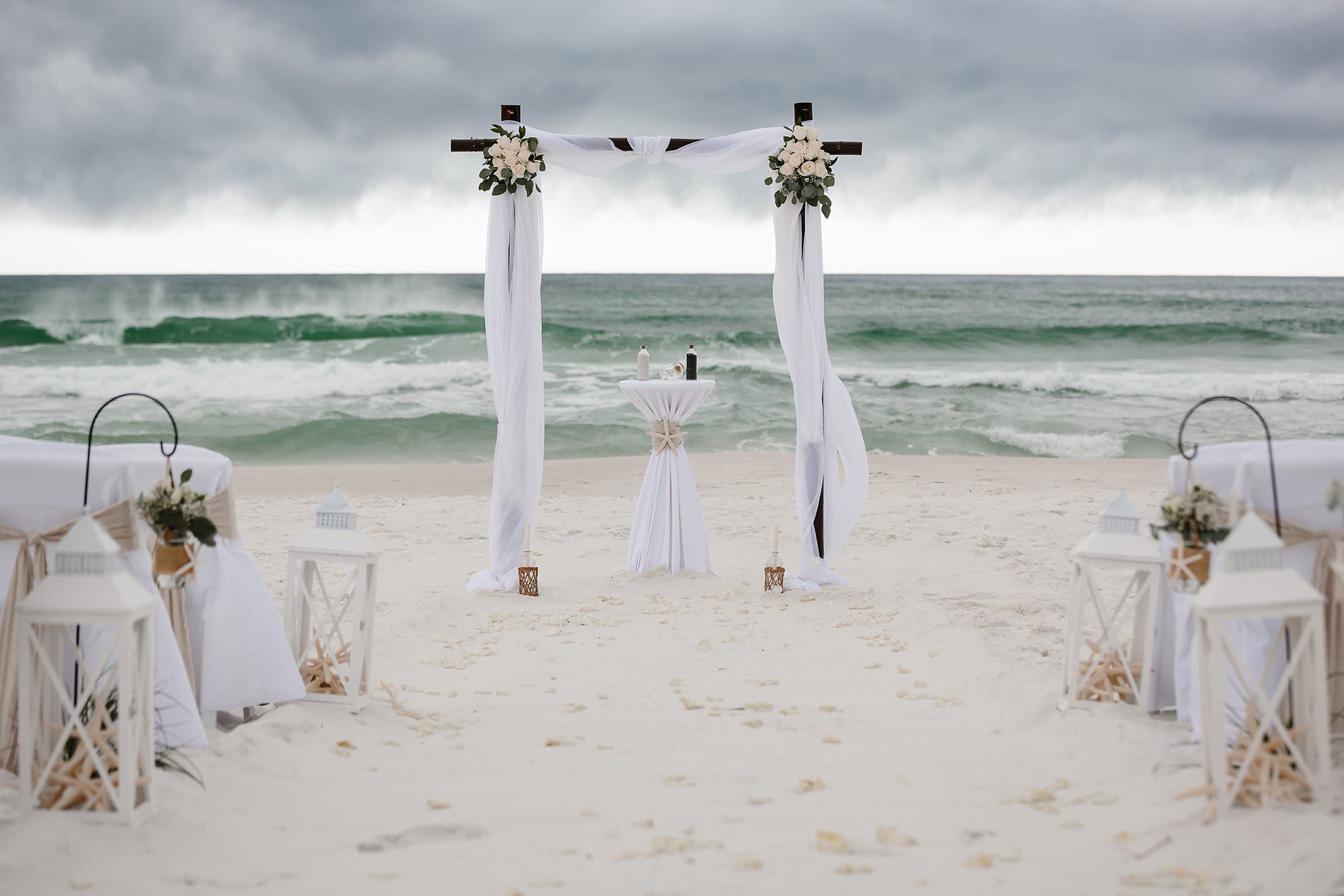 Beach wedding decor of bamboo arbor white roses lanterns white linen covered chairs facing dark storm clouds and white capped waves on Henderson Beach in Destin Florida
