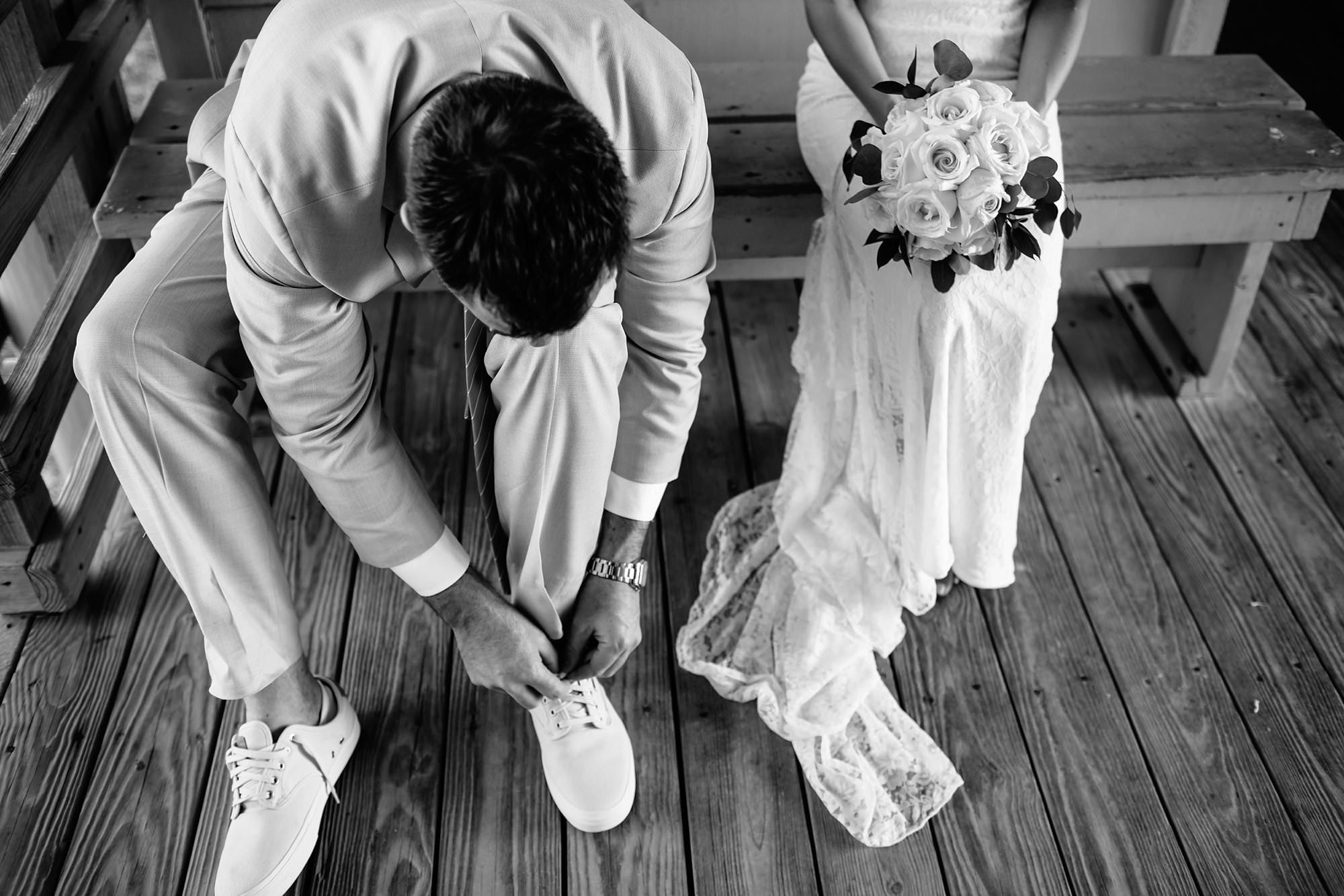 Black and white portraits of bride and groom sitting on beach while groom puts on shoes