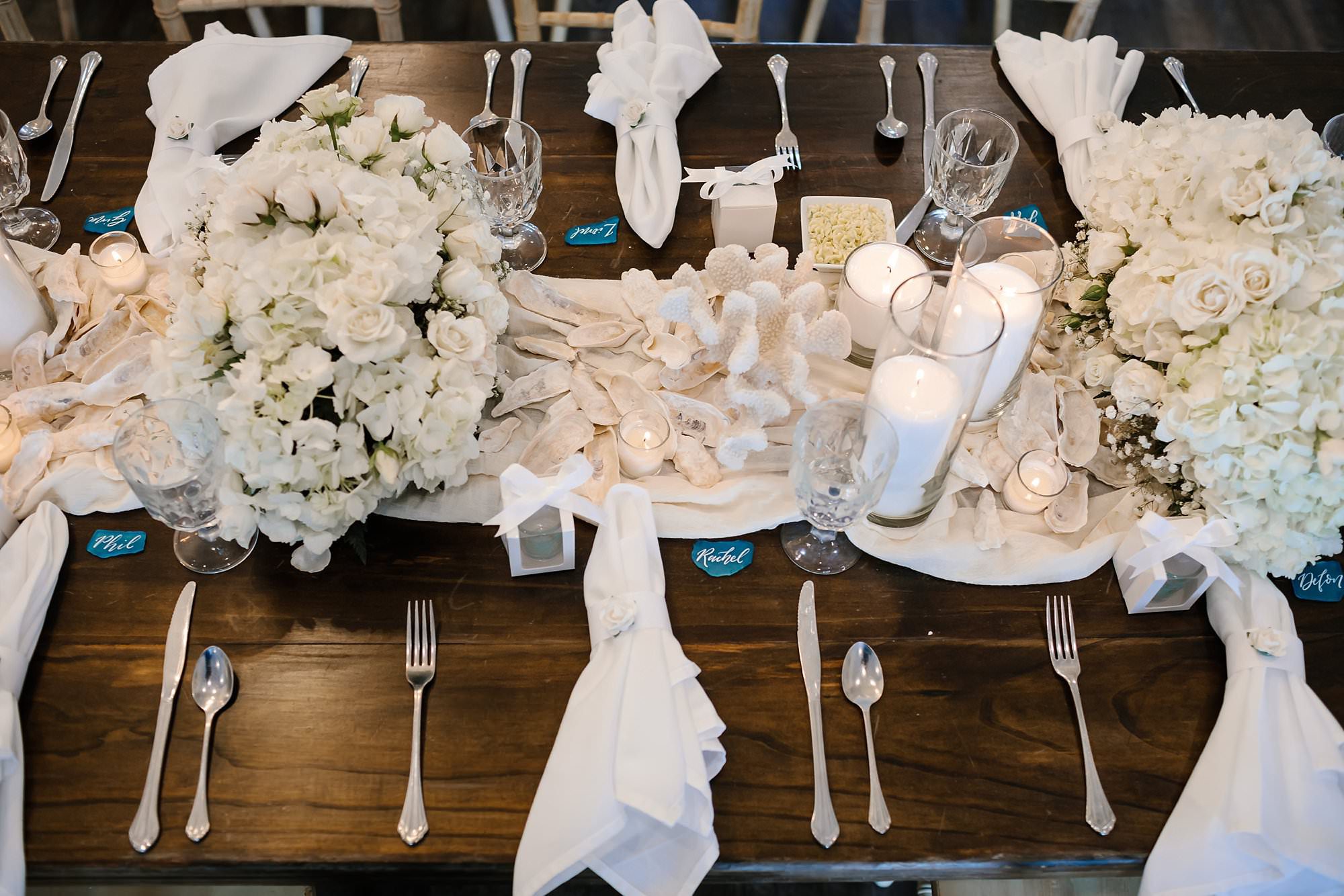 Lay flay of wedding reception table decor of dark wood farm table set with white rose centerpieces white shells candles white linen and aqua sea glass place cards