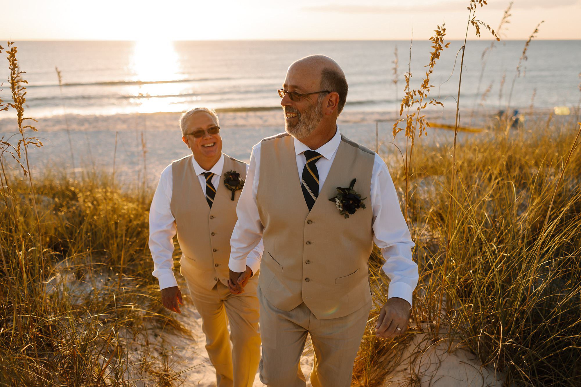 Sunset portrait of grooms walking hand and hand up a steep dune on the Gulf of Mexico