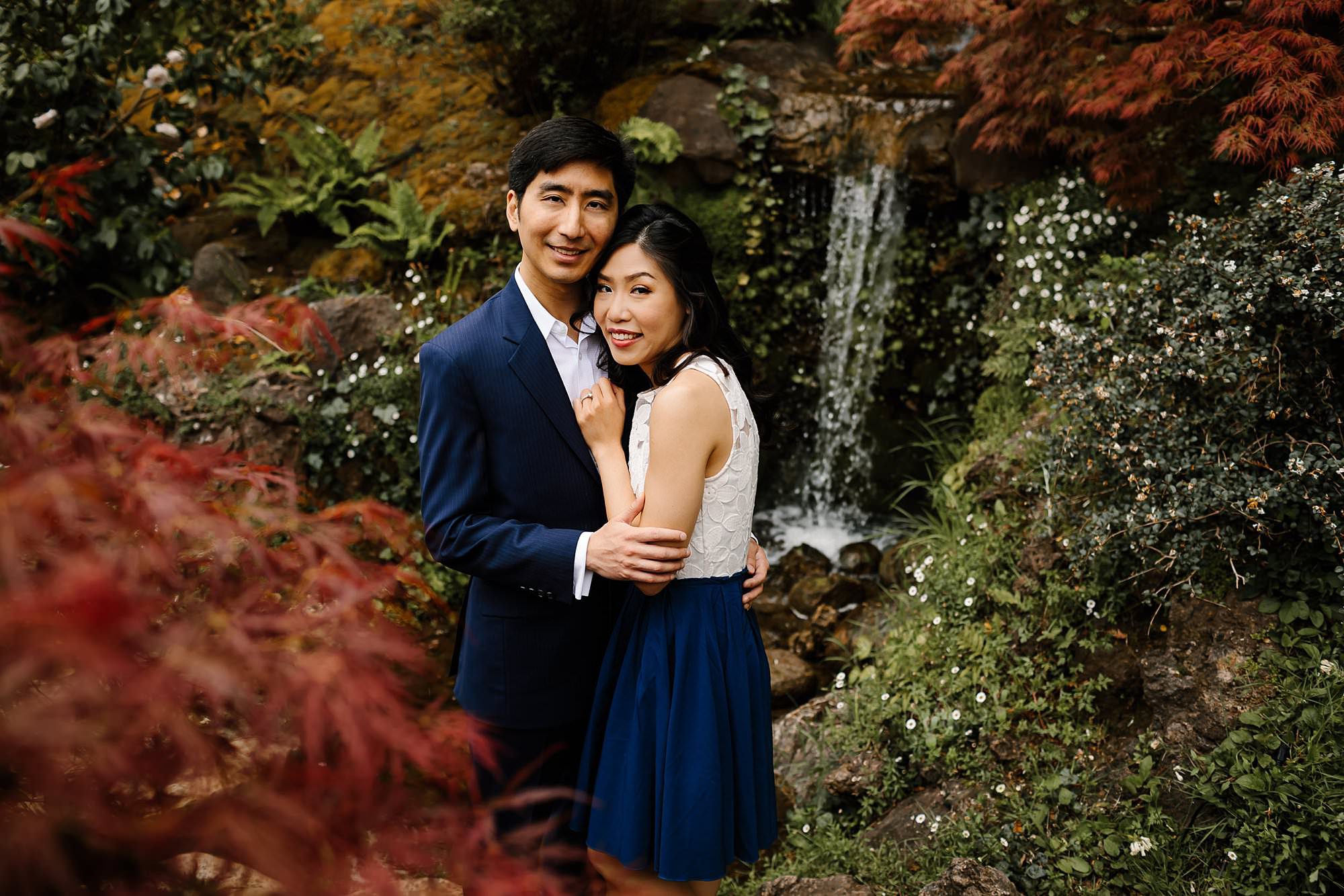 Engaged couple embracing at the waterfall surrounded by colorful foliage in Hakone Estate and Gardens in Saratoga California 