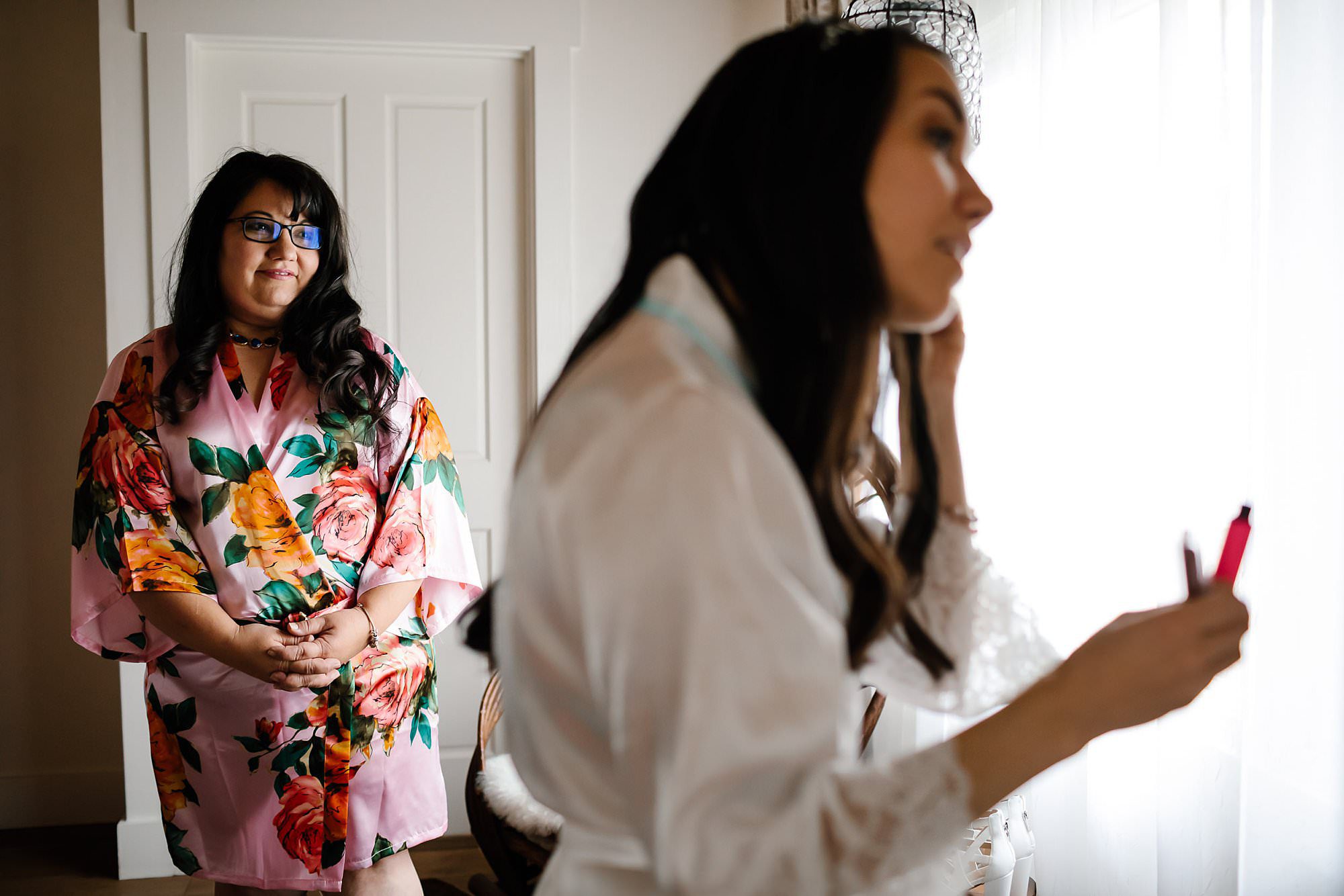 Mother of the bride in a floral robe watches bride apply mascara 